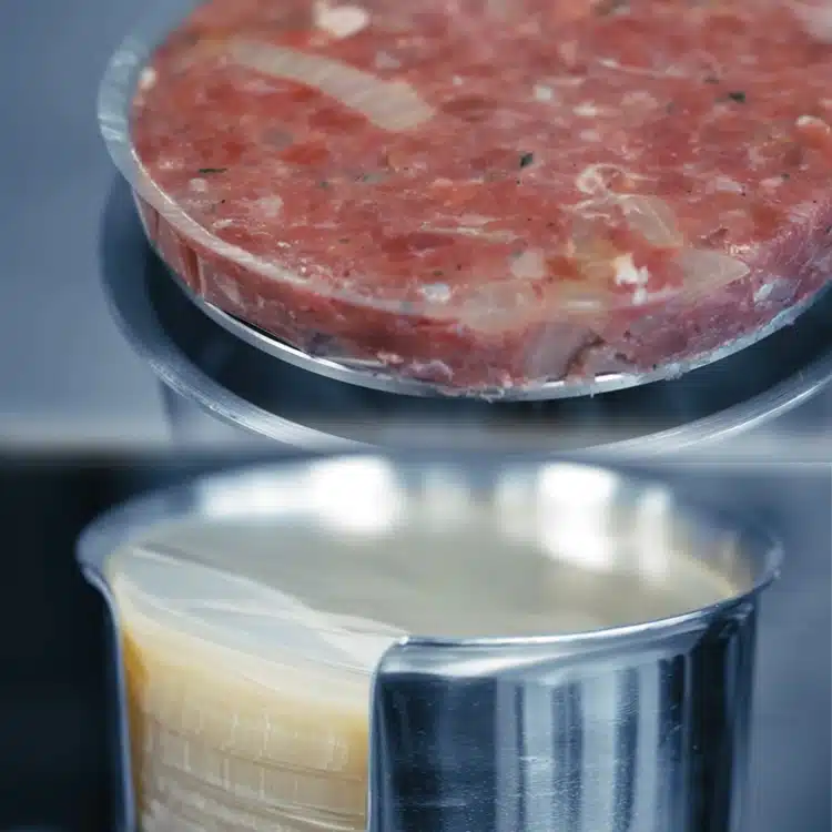 Raw burger patty on a cellophane patty disc, above a stainless steel holder loaded with patty discs